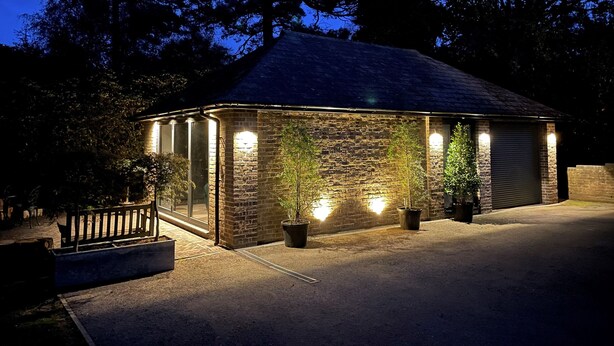 A stone building with a sloped roof, illuminated by exterior lights, surrounded by trees. - The Observatory Annexe (Dorking)