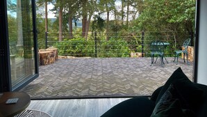 A room with a large sliding glass door leading to an outdoor patio with a herringbone-patterned floor, a wooden deck, and a view of a forested area. - The Observatory Annexe (Dorking)