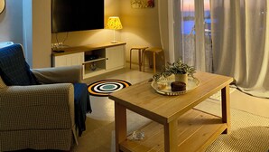 Un salon chaleureux avec un téléviseur à écran plat, une table basse en bois agrémentée d’une plante, un canapé à carreaux et un petit coin repas.
