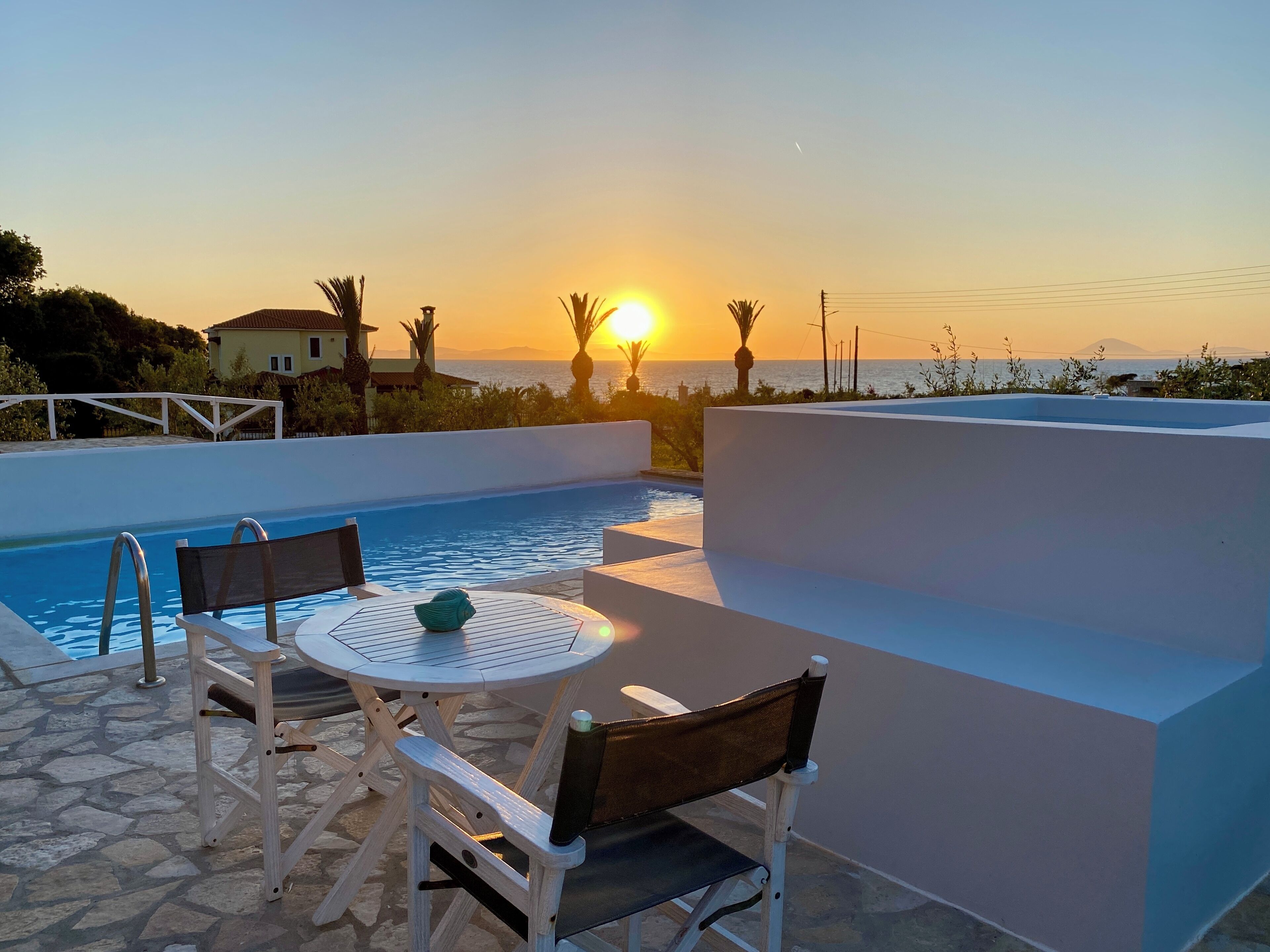 Área de piscina com mesa e cadeiras, com vista para o pôr do sol.