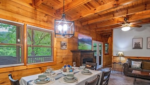 A wooden cabin interior with a dining table set for four, a fireplace, and a cozy living area with a couch and a television. - Location! Hot Tub, Game Room, Playground, 2K+ Sq Feet, Easy Roads! (Gatlinburg)