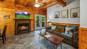 A cozy cabin interior with a fireplace, wooden walls, a sofa, and a television. - Location! Hot Tub, Game Room, Playground, 2K+ Sq Feet, Easy Roads! (Gatlinburg)