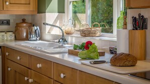 A kitchen with wooden cabinets, a sink, and various kitchen items like a loaf of bread, vegetables, and a knife block. - The Granary, Dunstall Green (Newmarket)