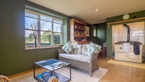 A cozy living room with a sofa, a glass coffee table, and a wicker chair. There's a window with a view of greenery outside. - The Granary, Dunstall Green (Newmarket)