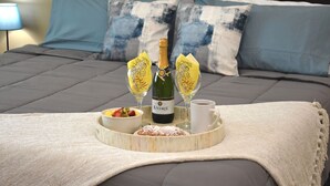 Un lit avec une couette grise, un plateau en bois avec une bouteille de vin, deux verres avec des étiquettes jaunes, un bol de fruits et une tasse de café.
