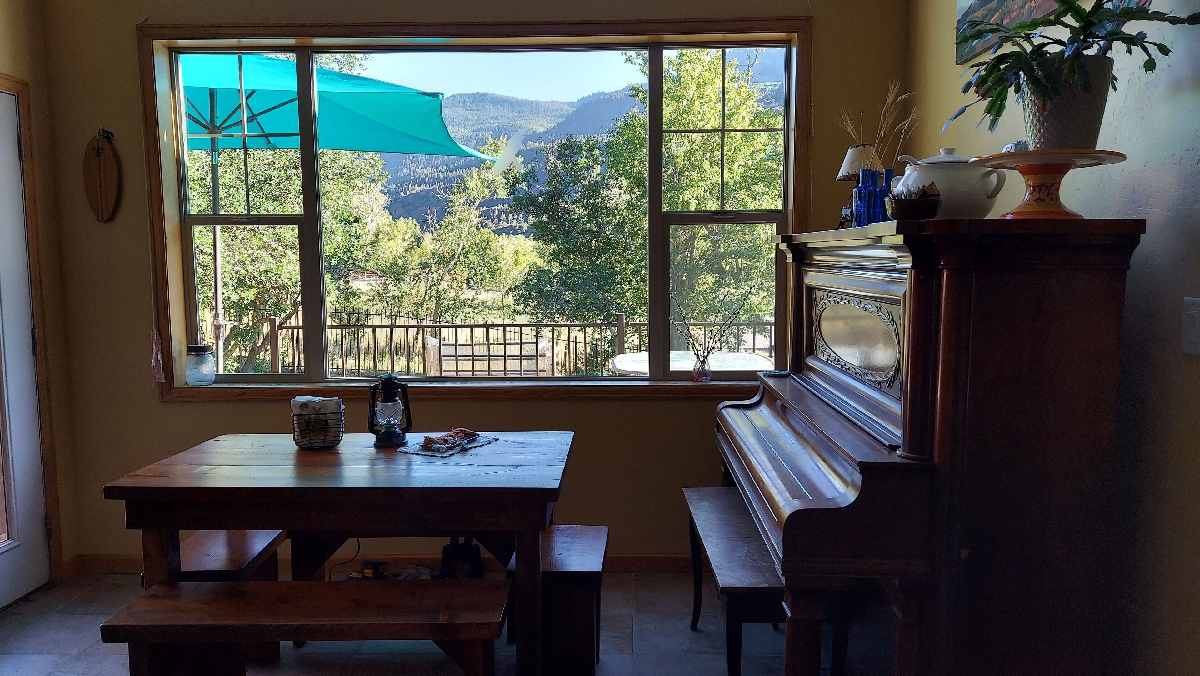A room with a large window offering a view of a forested area and mountains. There is a wooden table with a bench, a potted plant, and a decorative vase on a stand.