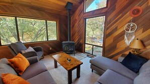 A cozy cabin interior with a sofa, armchair, and a small table. A wood-burning stove is visible, and there are large windows offering a view of trees. - Private house close to Sequoia National Park with pool and private river access (Three Rivers)