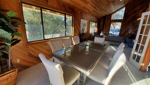 A dining area with a glass table and white upholstered chairs, wooden walls, and large windows overlooking a garden. - Private house close to Sequoia National Park with pool and private river access (Three Rivers)