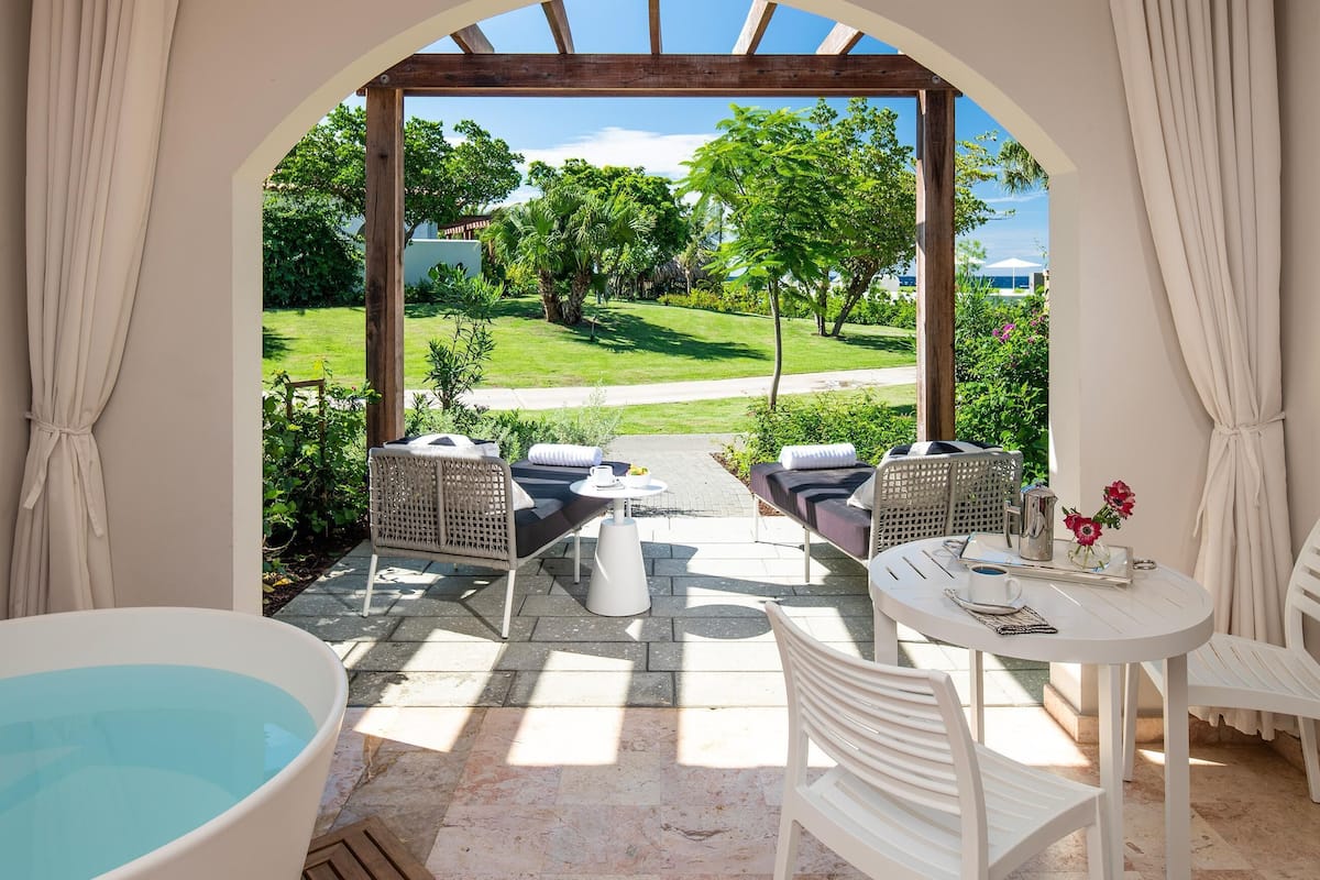 Ein Wellnessbereich mit einer Badewanne, zwei Korbstühlen, einem kleinen Tisch und Blick auf einen Garten.