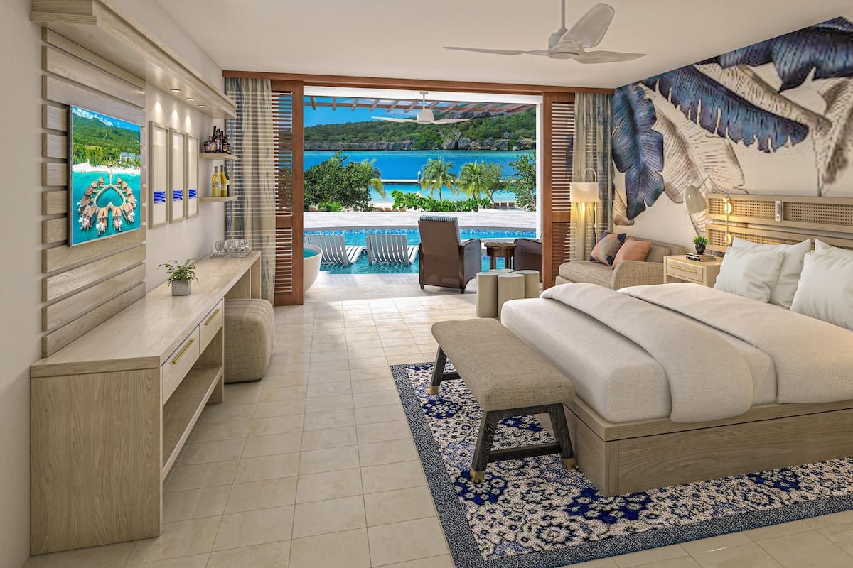 Ein Schlafzimmer mit einem großen Bett, einem Nachttisch, einem Schreibtisch, einem Stuhl, einer Bank und Blick auf Pool und Strand.