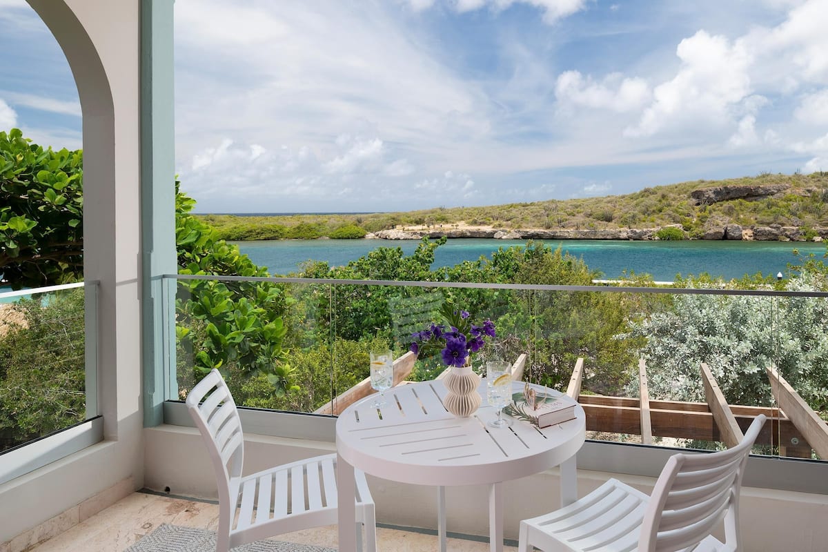 Ein Balkon mit Tisch und Stühlen mit Blick auf ein türkisblaues Meer und eine grüne Landschaft.