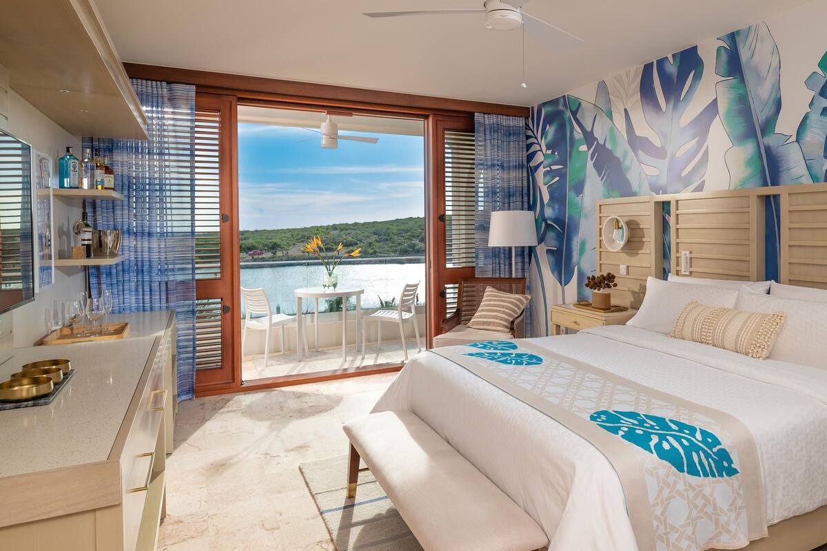 Ein Schlafzimmer mit einem großen Bett, einem Schreibtisch mit Stuhl, einem Balkon mit Tisch und Stühlen sowie Blick auf das Wasser.