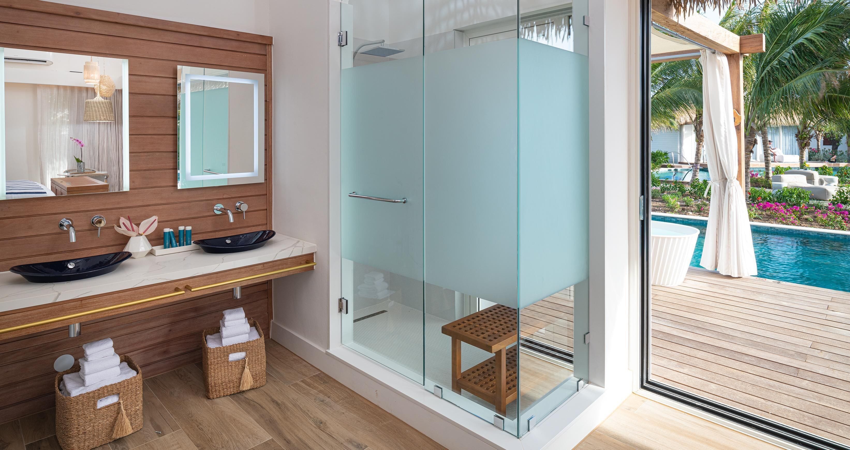 A modern bathroom with a glass-enclosed shower, a wooden vanity with a large mirror, and a view of a pool outside.
