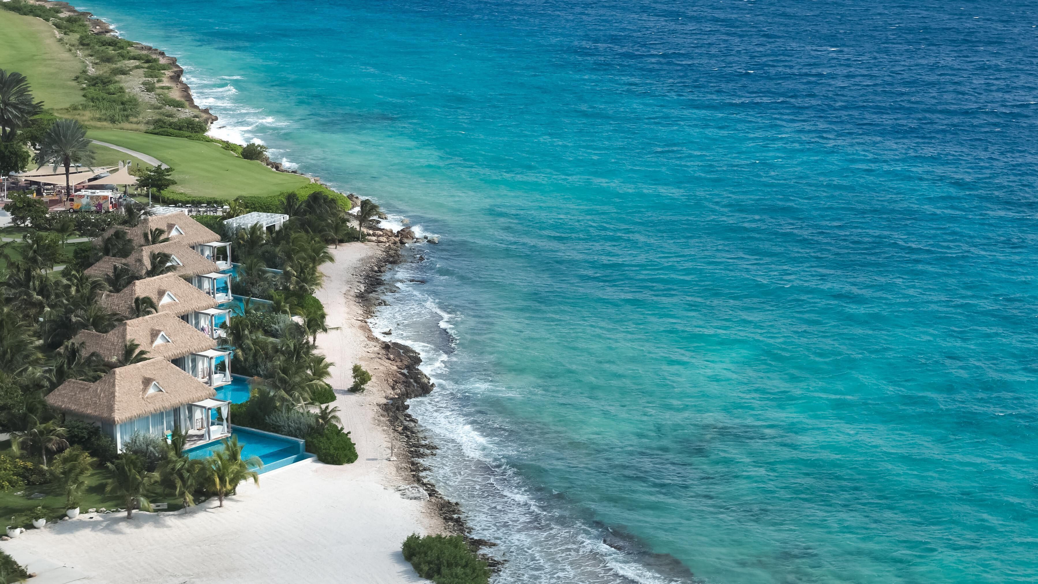An aerial view of a coastal resort with multiple bungalow-style accommodations, a sandy beach, and a clear blue ocean.