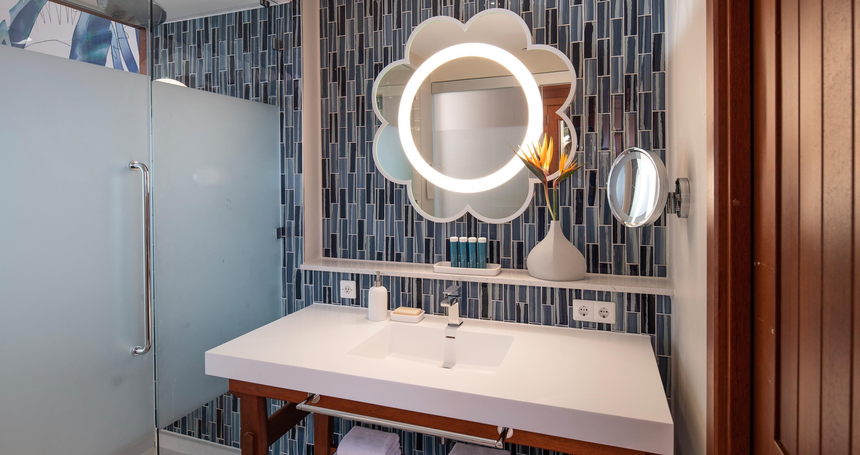 A bathroom with a white sink, a mirror with a circular light, and a blue patterned wall.