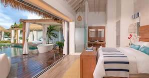 A modern hotel room with a wooden deck, a large bed, a bathroom with a vanity, and a view of a pool area.