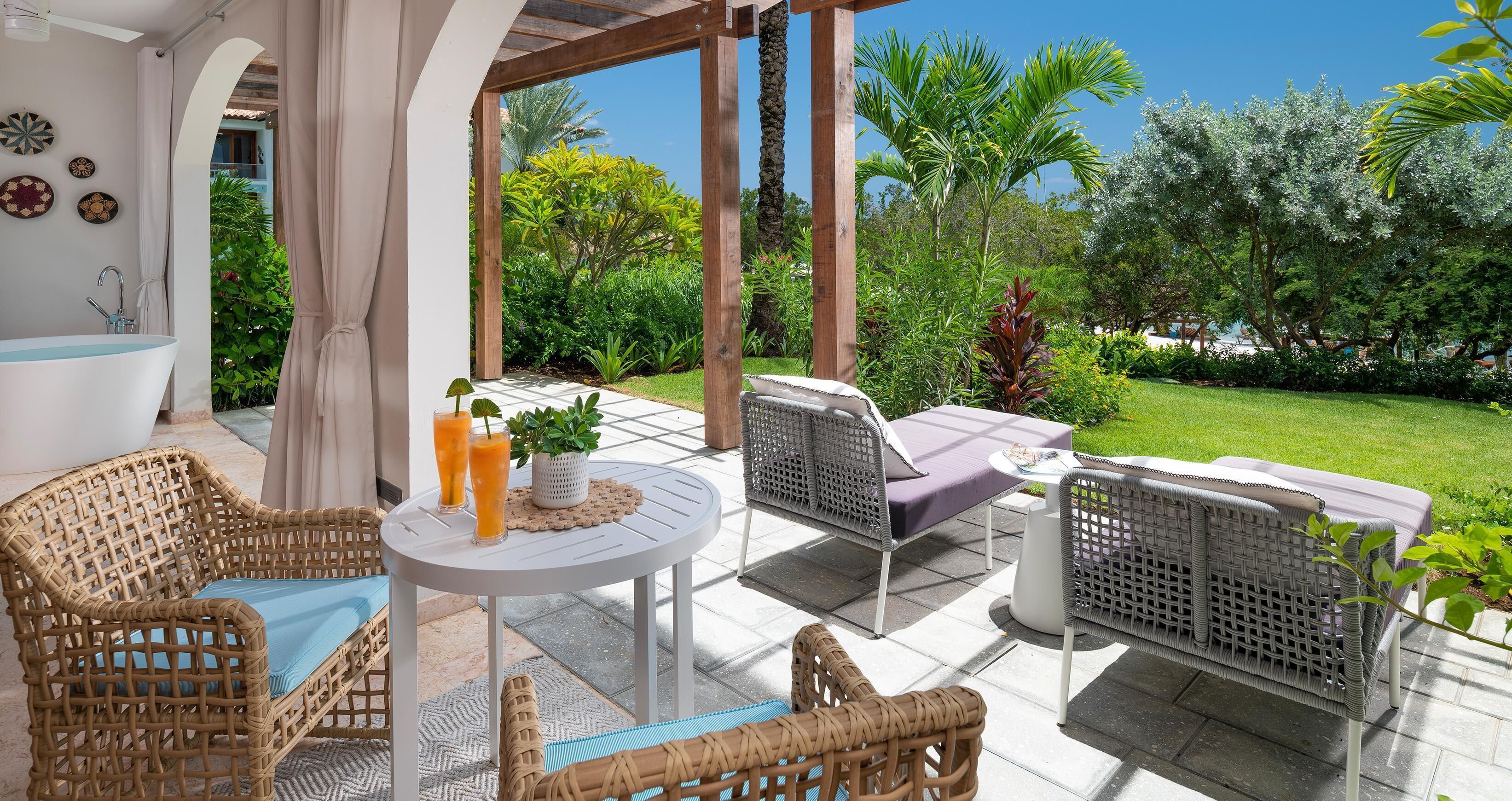 A covered patio area with wicker furniture, a round table, and a view of a lush garden.