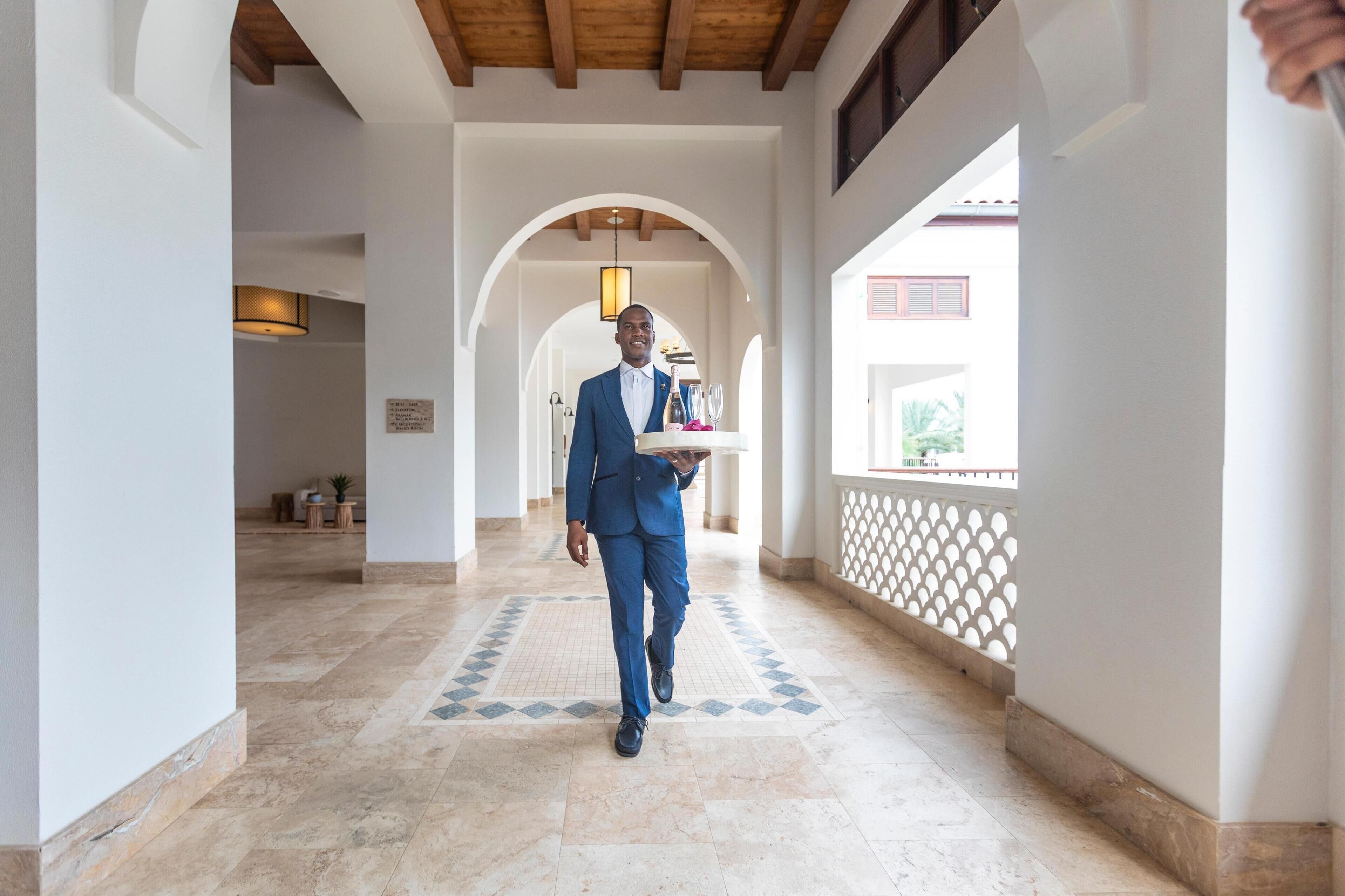 A man in a blue suit walking down a hallway with a tray of items.