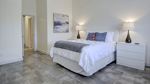 Un dormitorio moderno con una cama grande, dos mesitas de noche con lámparas, una mesita auxiliar, un cuadro colgado en la pared y un pasillo que conduce a otra habitación.