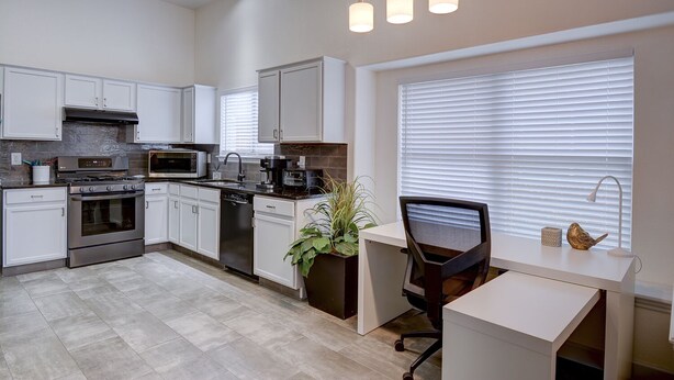 Una cocina moderna con gabinetes blancos, electrodomésticos de acero inoxidable, horno empotrado y un comedor con mesa y silla blancos.