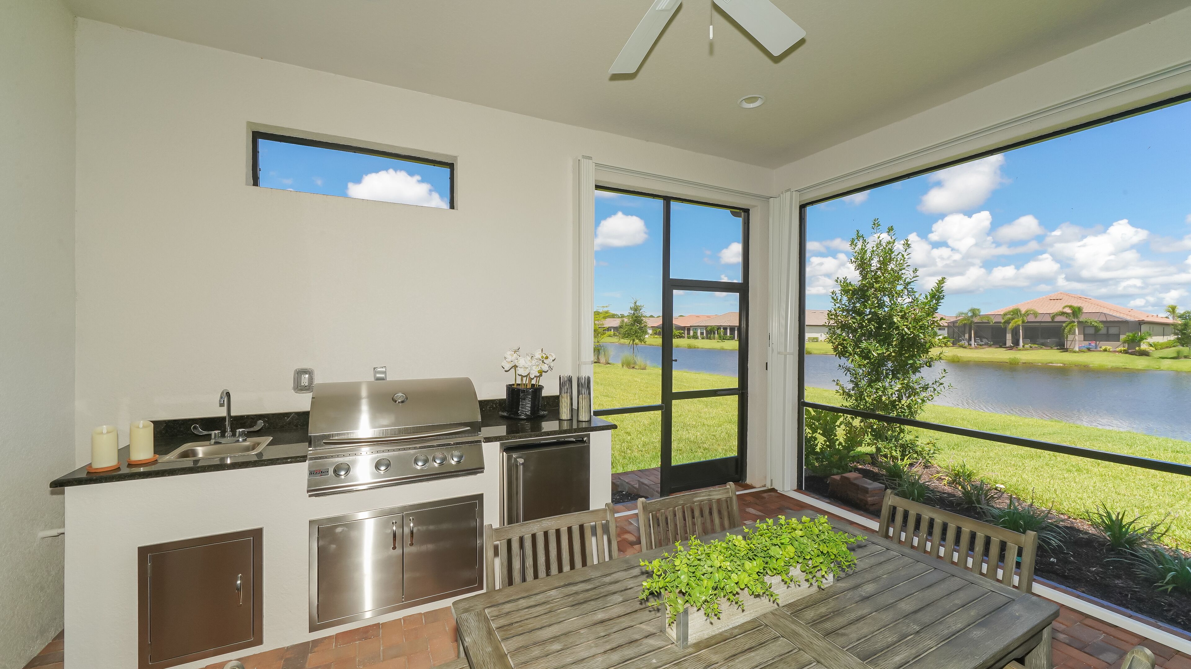 A modern kitchen with a built-in grill, a dining area with a table and chairs, and large windows offering a view of a lake and houses.