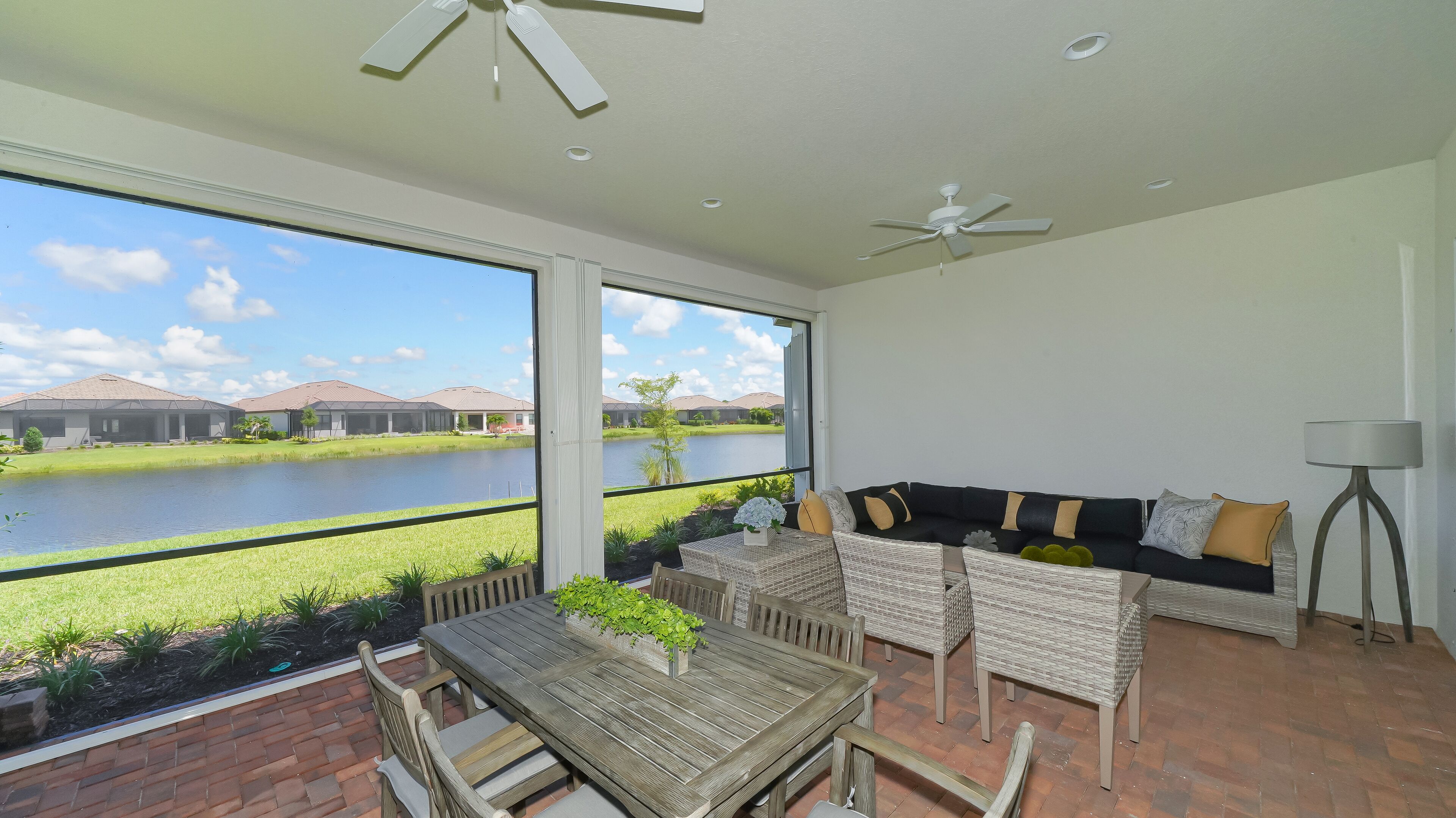 A spacious balcony with a view of a lake and houses, furnished with a wooden table, wicker chairs, and a black sofa with cushions.