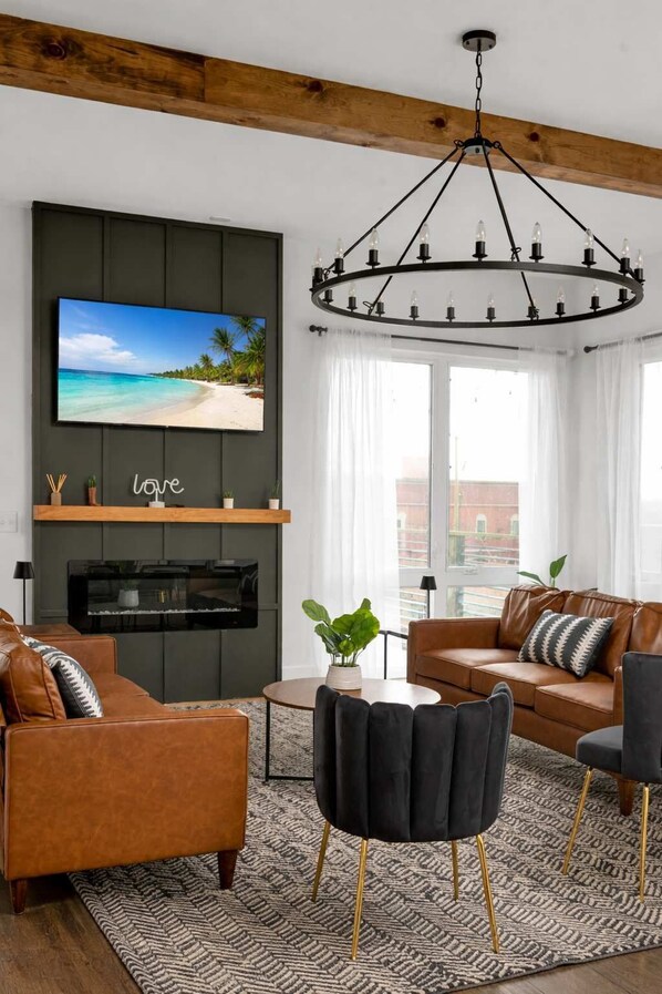 A modern living room with a TV mounted on a dark wall, a chandelier, leather sofas, a coffee table, and a patterned rug.