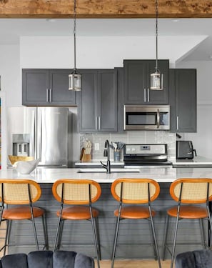 Una cocina moderna con gabinetes grises, electrodomésticos de acero inoxidable y una encimera blanca. Hay cuatro taburetes de bar con asientos naranjas y estructura de metal negro.