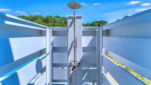 Une douche extérieure moderne, dotée d’une pomme de douche circulaire, entourée de murs blancs et surplombée par un ciel bleu clair.