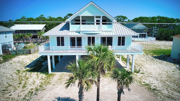 Une maison de plage à deux étages, dotée d’un balcon, entourée de sable et de palmiers.