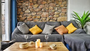 A cozy living room with a stone wall, a grey sofa with cushions, a wooden coffee table with candles and a glass, and a potted plant.