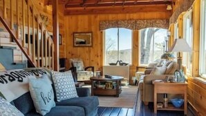 L’intérieur d’un chalet accueillant, avec un escalier en bois, un canapé, une table à manger et une fenêtre donnant sur les arbres.