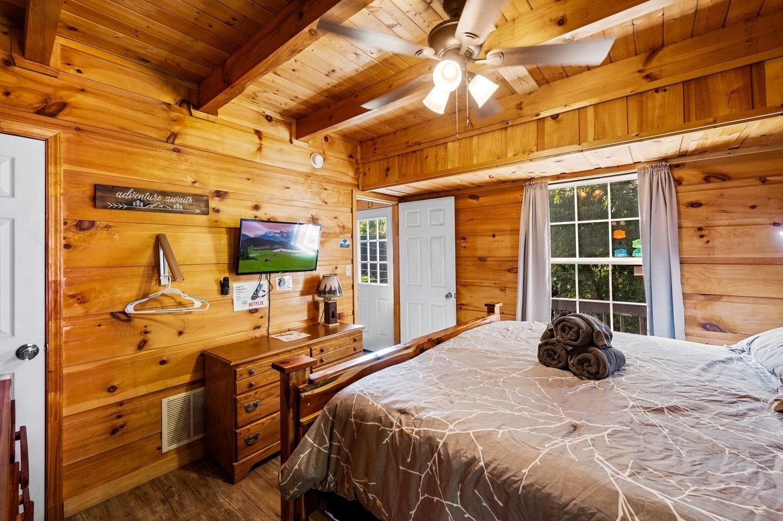 Un dormitorio de cabaña de madera con una cama, un televisor, un aparador y un ventilador de techo.