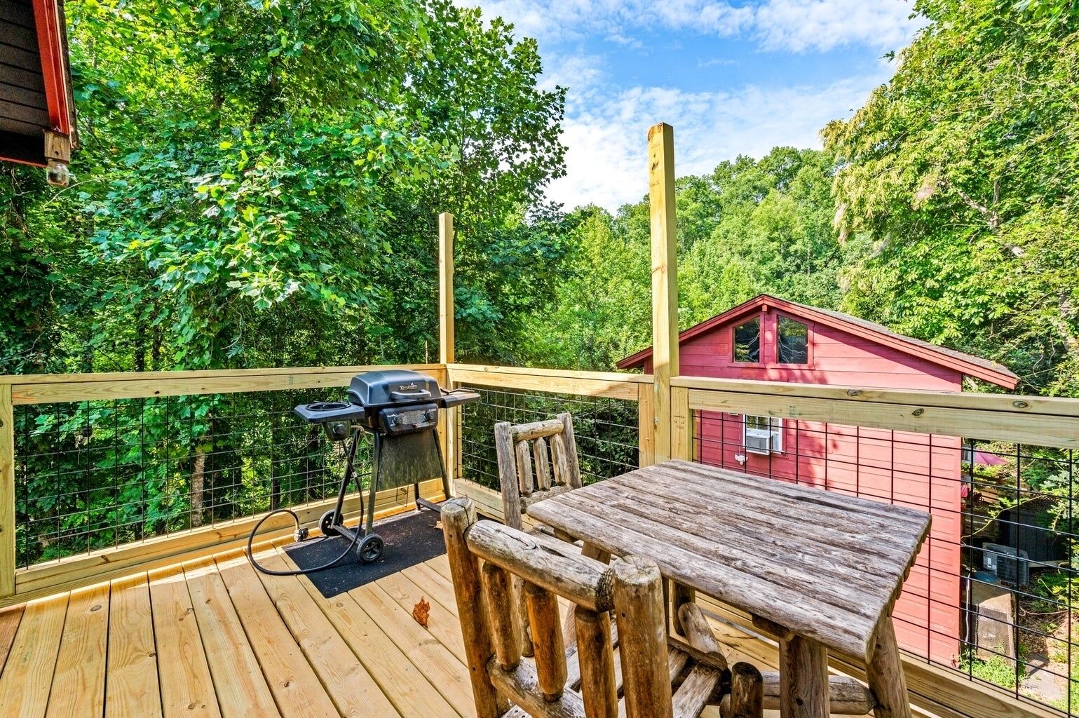 Een houten terras met een barbecue, een tafel en stoelen, omgeven door een rood gebouw en weelderige begroeiing.