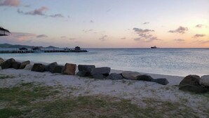 Una playa con costa rocosa, un muelle que se adentra en el agua y un barco a lo lejos.