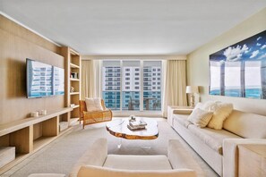 A modern hotel room with a large window, a flat-screen TV, a comfortable sofa, a wicker chair, and a coffee table.