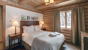 A wooden cabin bedroom with a bed, two bedside lamps, a nightstand, a window with curtains, and framed artwork on the wall. - Stunning Log Chalet - Private Hot Tub, Pool Table, Fireplace (Labelle)