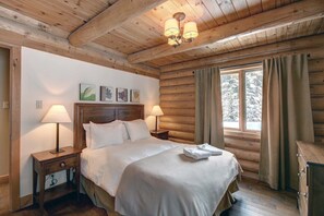 Ett sovrum i en stuga med träpanel, inrett med en säng, två sänglampor, ett nattduksbord, ett fönster med gardiner och inramade konstverk på väggen.