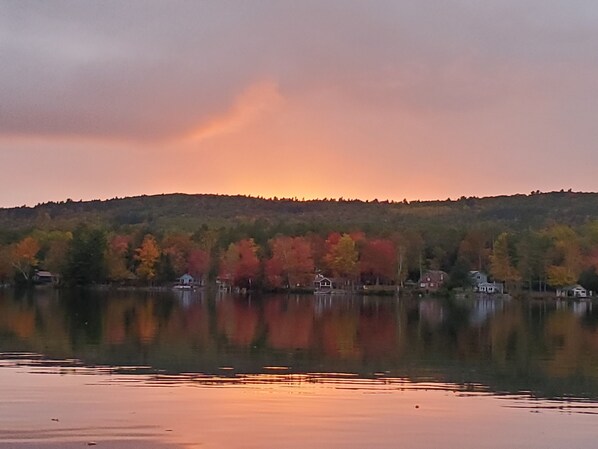 Un lac paisible, bordé d’arbres aux couleurs automnales qui se reflètent dans l’eau, et entouré d’une colline boisée.