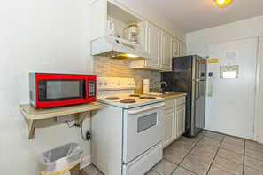 A compact kitchen with a microwave, stove, refrigerator, and a small trash bin.