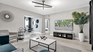 A modern living room with a sofa, coffee table, and TV. There is a dining area with a table and chairs, and a large plant in the corner. - Madison Suite 2 BD/2BA Miami Beach Lincoln Casa (Miami Beach)