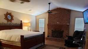 A bedroom with a large bed, a brick fireplace, a ceiling fan, and a television on a stand.