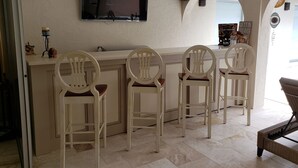 A bar area with four white chairs, a flat-screen TV mounted on the wall, and a small table.