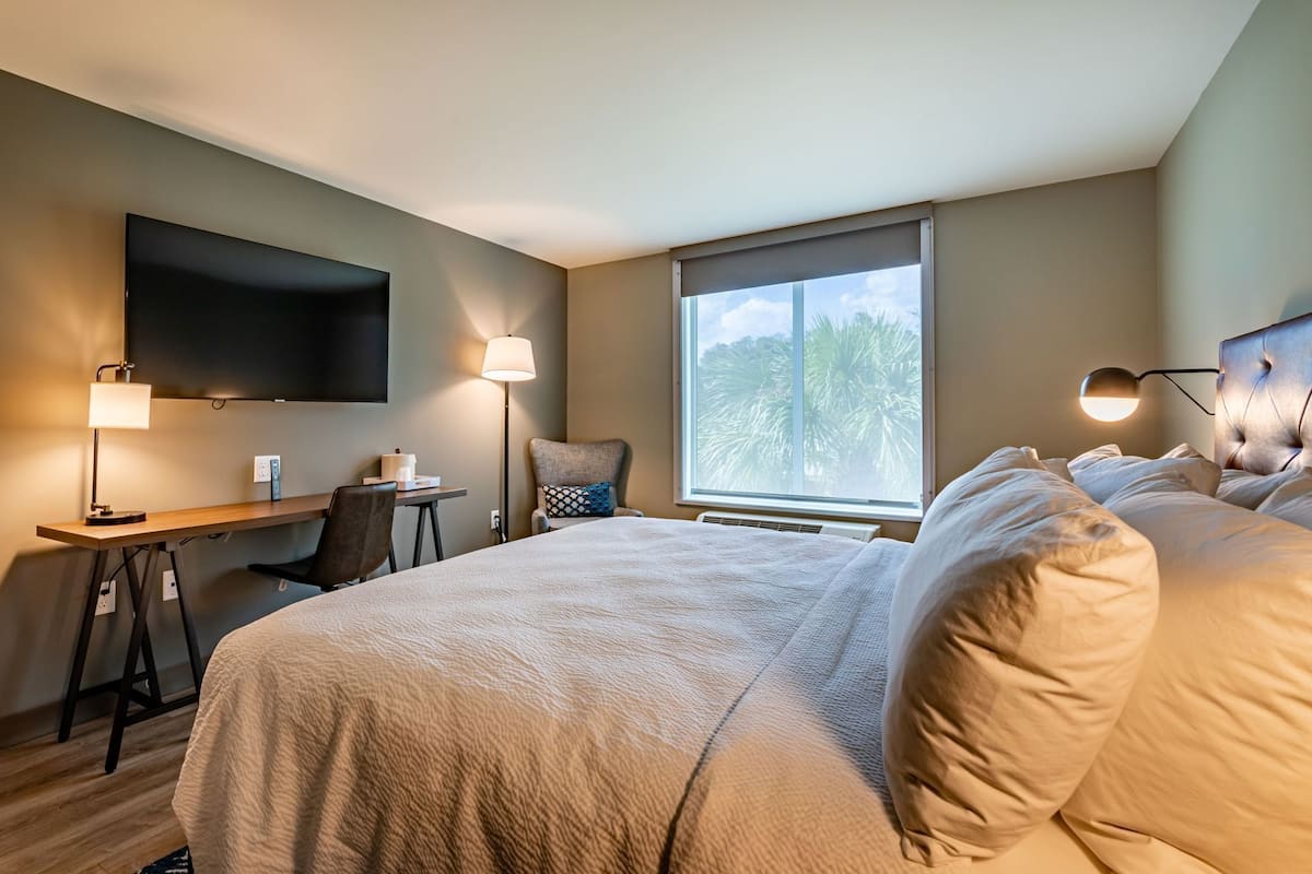 Habitación de hotel con una cama grande, un escritorio, una silla, un televisor montado en la pared y una ventana con vistas a árboles.