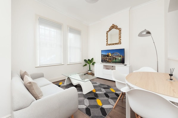 A modern living room with a sofa, a glass coffee table, a dining table, a TV, and a plant.