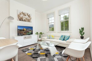 Un salón moderno con televisor de pantalla plana, muebles blancos, una mesa de centro de vidrio y vistas a zonas verdes.