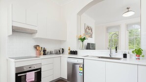 Une cuisine moderne avec des meubles blancs, un four encastré, un lave-vaisselle et une grande fenêtre qui laisse entrer la lumière naturelle.