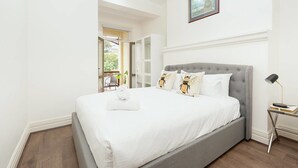Une chambre avec un lit recouvert d’un tissu gris, du linge de lit blanc et des coussins décoratifs ornés d’un motif d’abeille. Un tableau encadré est accroché au mur au-dessus du lit, une table de chevet avec une lampe s’y trouve, et on peut admirer un balcon extérieur depuis la chambre.