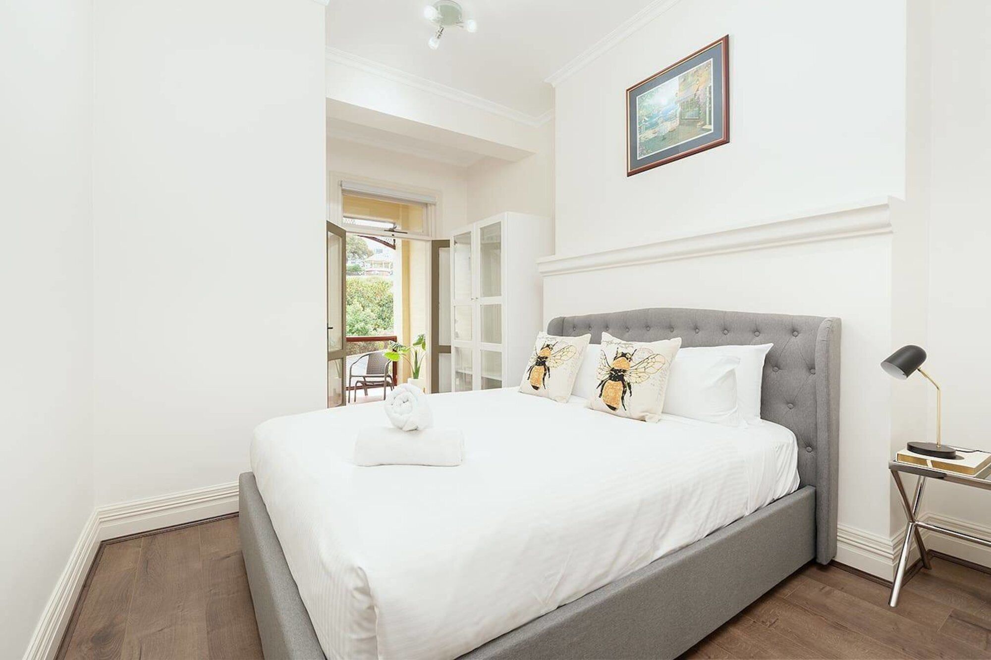 A bedroom with a grey upholstered bed, white bedding, and a painting on the wall.