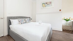 A neatly made bed with a grey headboard, white bedding, and a floral pillow. A rolled towel is placed at the foot of the bed. The room has a white wall with a framed abstract painting above the bed. - Stunning Sydney Home 8 (Millers Point)